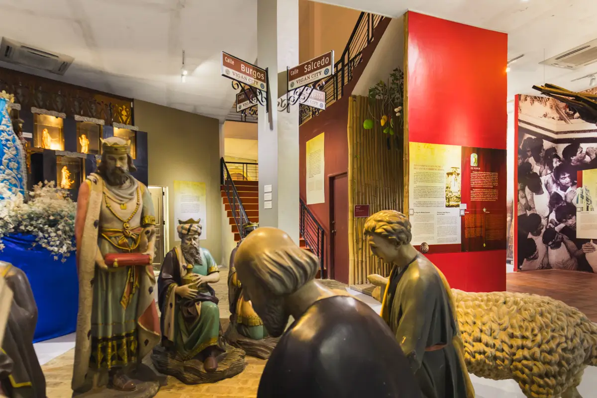 This museum exhibit, one of the best places in Vigan, features life-sized statues in biblical attire set against red and beige walls with text panels, a staircase, and directional signs—a must-see on any Vigan travel guide.