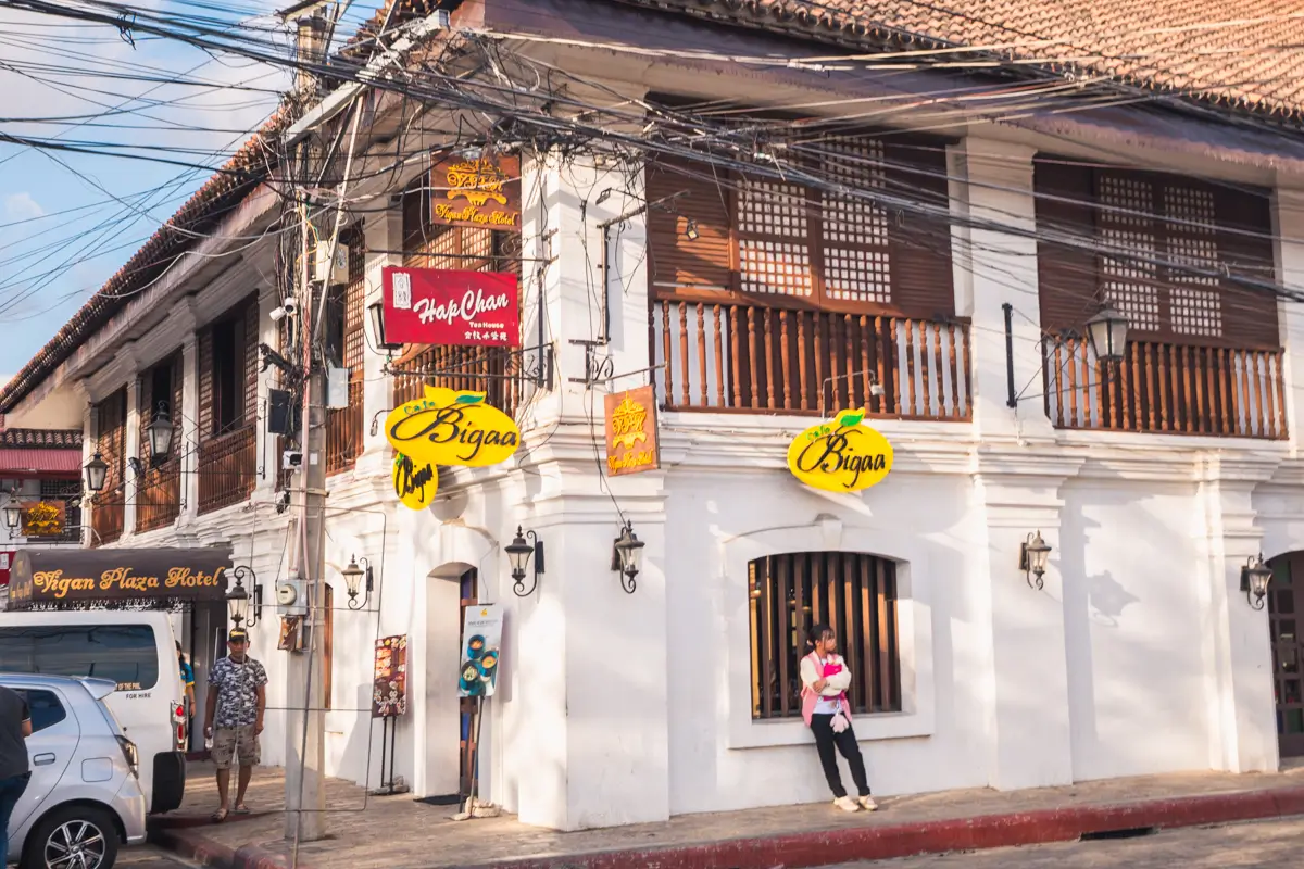 A woman leans against the exterior wall of a white colonial-style building, one of the best places in Vigan, with wooden windows and colorful signs—capturing the charm found in many popular Vigan tourist spots.