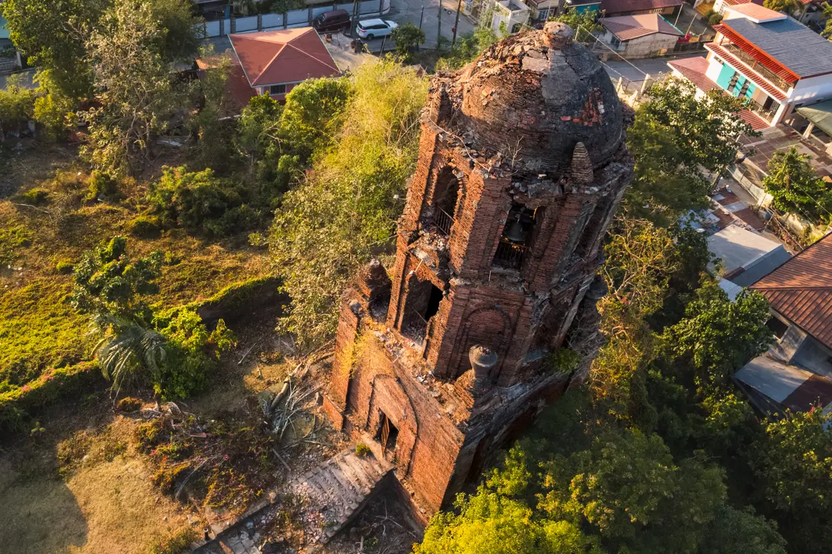 Aerial view of an old, crumbling brick bell tower surrounded by trees and nearby houses—one of the fascinating Vigan tourist spots, with the structure showing significant damage and missing roof sections.