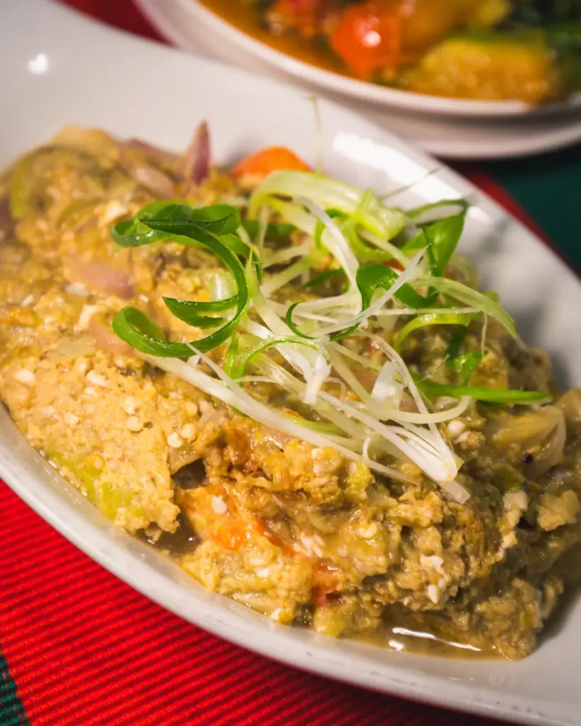 A close-up of a white dish filled with a beige, chunky vegetable and tofu stew, topped with sliced green onions—a must-try for those exploring best places in Vigan—with another plate of food blurred in the background.