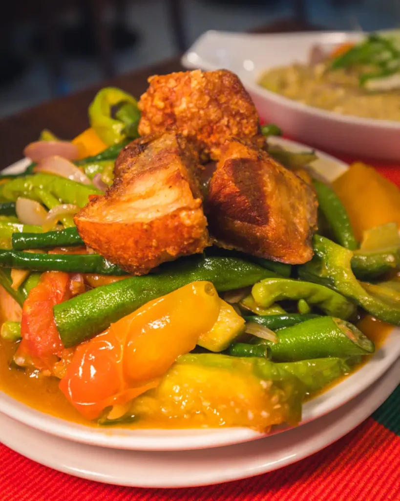 A plate of mixed vegetables with green beans and tomatoes, topped with crispy fried pork, served on a white dish—a must-try for anyone exploring Vigan tourist spots. Another delicious plate is slightly blurred in the background.