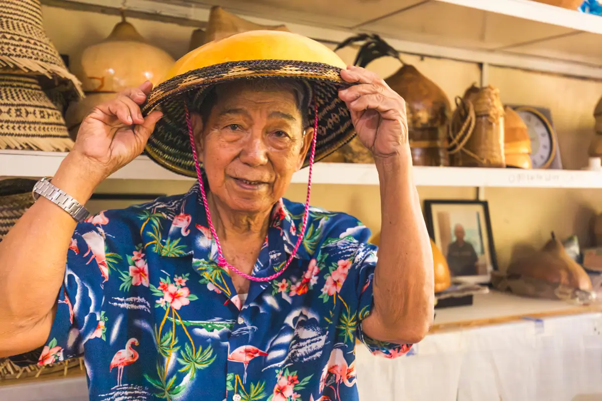 An older person wearing a colorful floral shirt stands indoors, holding a woven hat on their head. Shelves behind them display traditional crafts, a clock, and framed photos—capturing the charm often highlighted in Vigan travel guides.