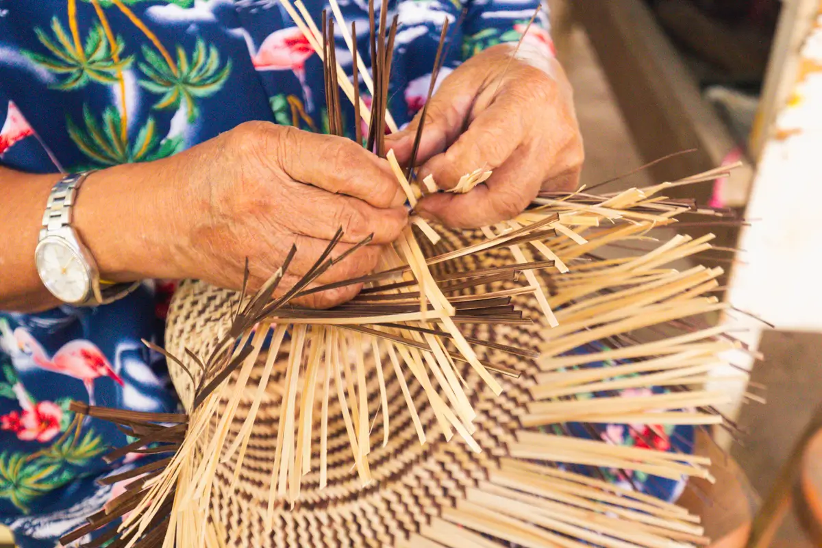 A person wearing a watch weaves light and dark reeds into a patterned basket, dressed in a colorful shirt with floral and flamingo designs—capturing the artistry you’ll find at the best places in Vigan.