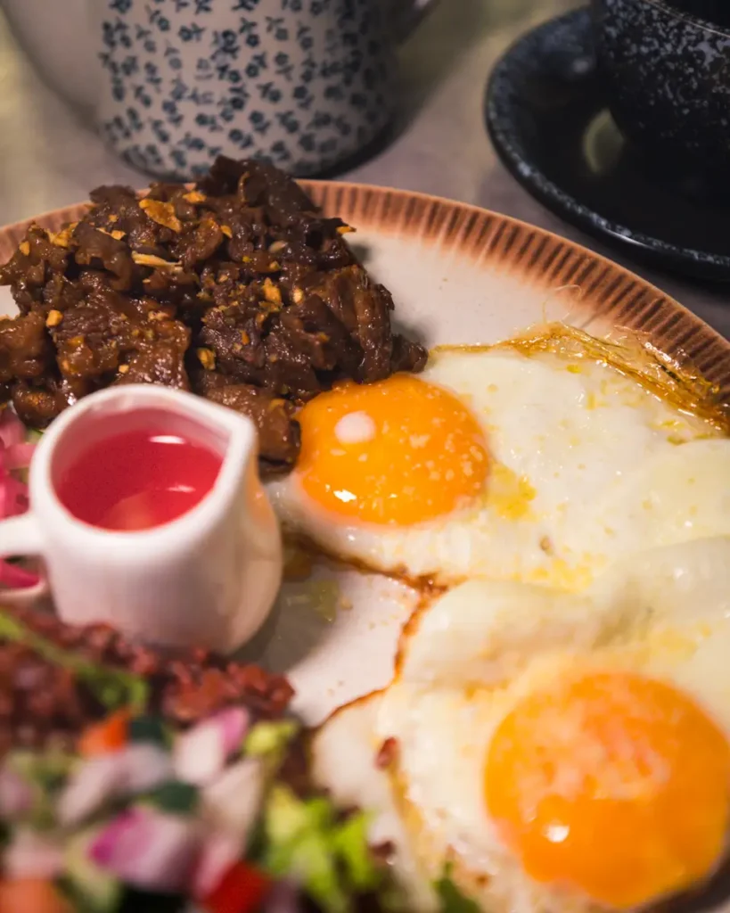 A plate with two sunny-side up eggs, a portion of cooked meat or stew, a small pitcher of red sauce, and a colorful vegetable salad—a taste of local cuisine often enjoyed when exploring the best places in Vigan. A mug and cup are partially visible in the background.