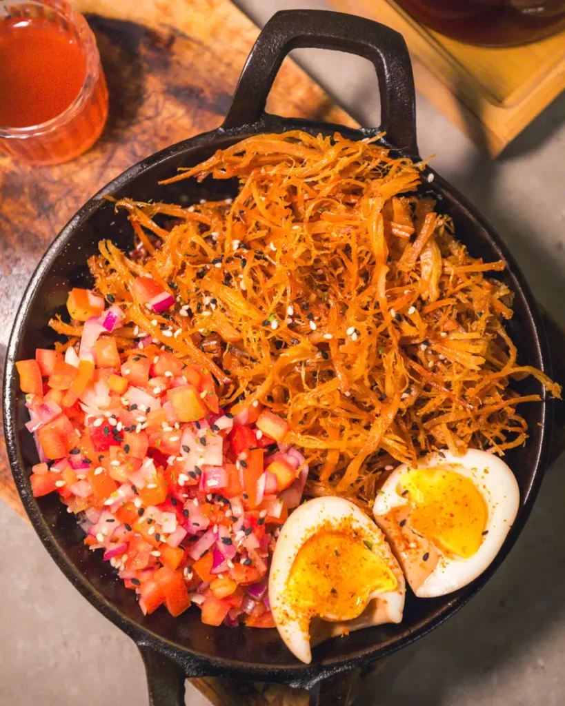 A black skillet holds shredded dried meat, a tomato and onion mix with sesame seeds, and a halved soft-boiled egg. This savory dish is a must-try for food lovers exploring Vigan Tourist Spots—a true highlight on any Vigan Guide.