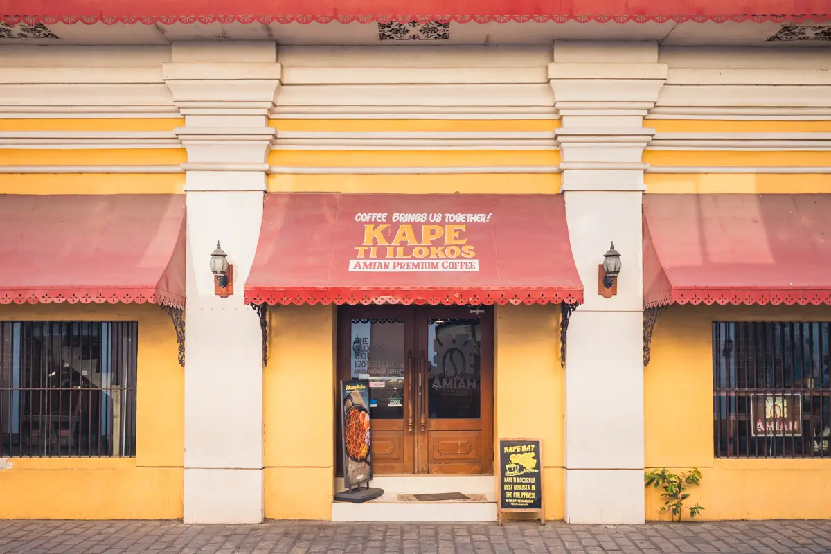 A yellow and white building with large columns features a red awning reading “Kape Ti Ilokos Amian Premium Coffee,” one of the best places in Vigan. Two lanterns flank a wooden door, with signs for food and drinks displayed outside the entrance.