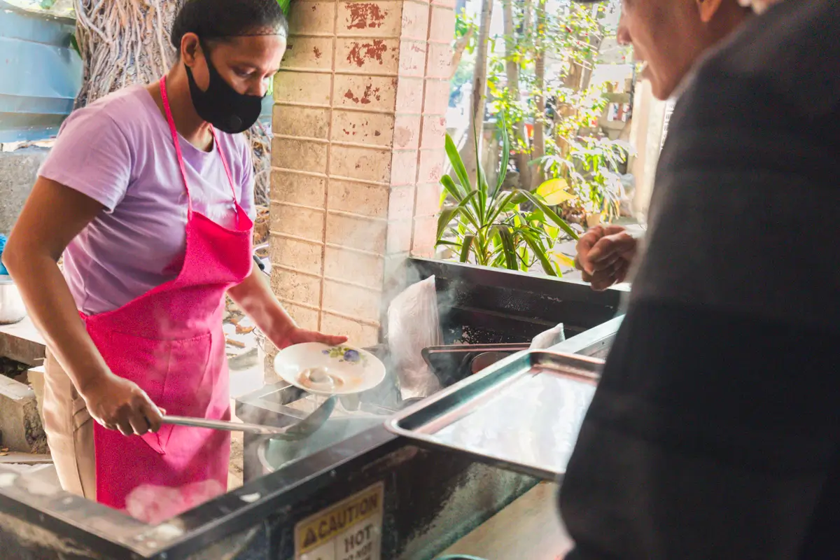 A woman in a pink apron and black face mask serves food onto a white plate at a food stall, while a customer waits across the counter—a scene you might find while exploring the best places in Vigan. Steam rises from the busy cooking area.