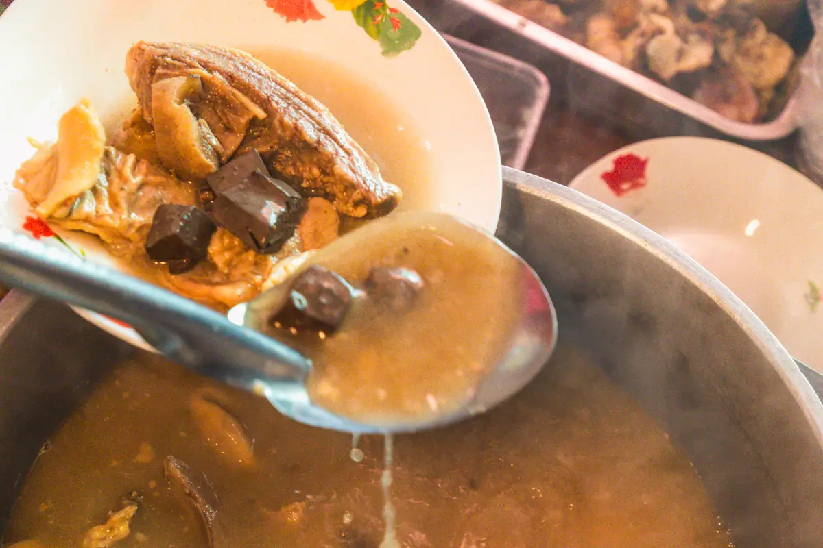 A close-up of a bowl being filled with soup from a large pot, capturing the flavors that make up some of the Best Vigan dishes. Pieces of meat, brown cubes, and broth are ladled in as steam rises—a treat for any Vigan guide to local cuisine.