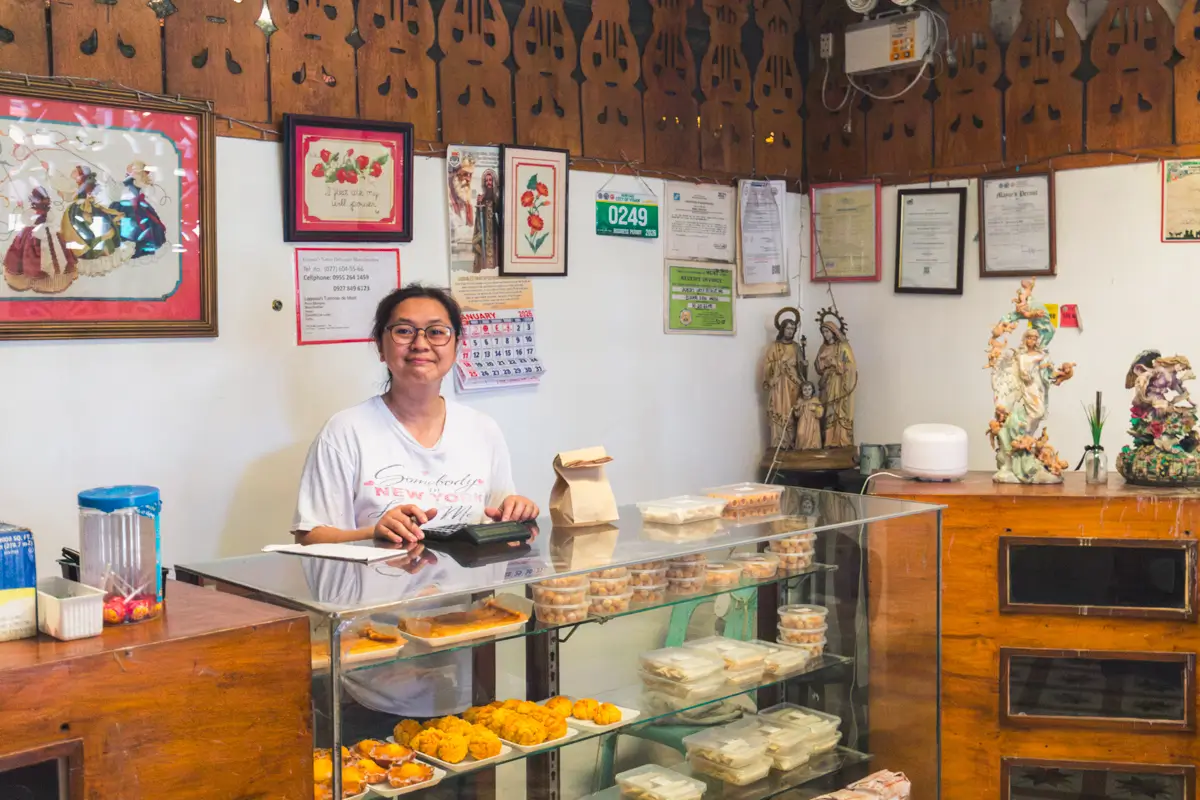 A woman stands behind a glass display case filled with baked goods in a charming bakery—one of the best places in Vigan. The walls feature framed art, certificates, and decorations; the counter is topped with a cash register, snacks, and packaged foods.