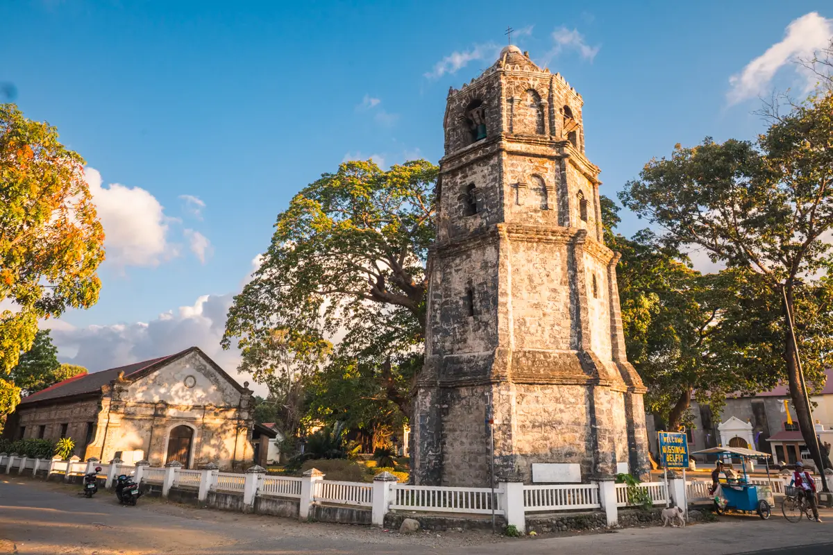 A tall stone bell tower stands next to a small church building, surrounded by large trees and a white fence, under a clear blue sky with clouds—a charming scene often featured in Vigan tourism guides among top Vigan tourist spots. Vehicles are parked along the street.