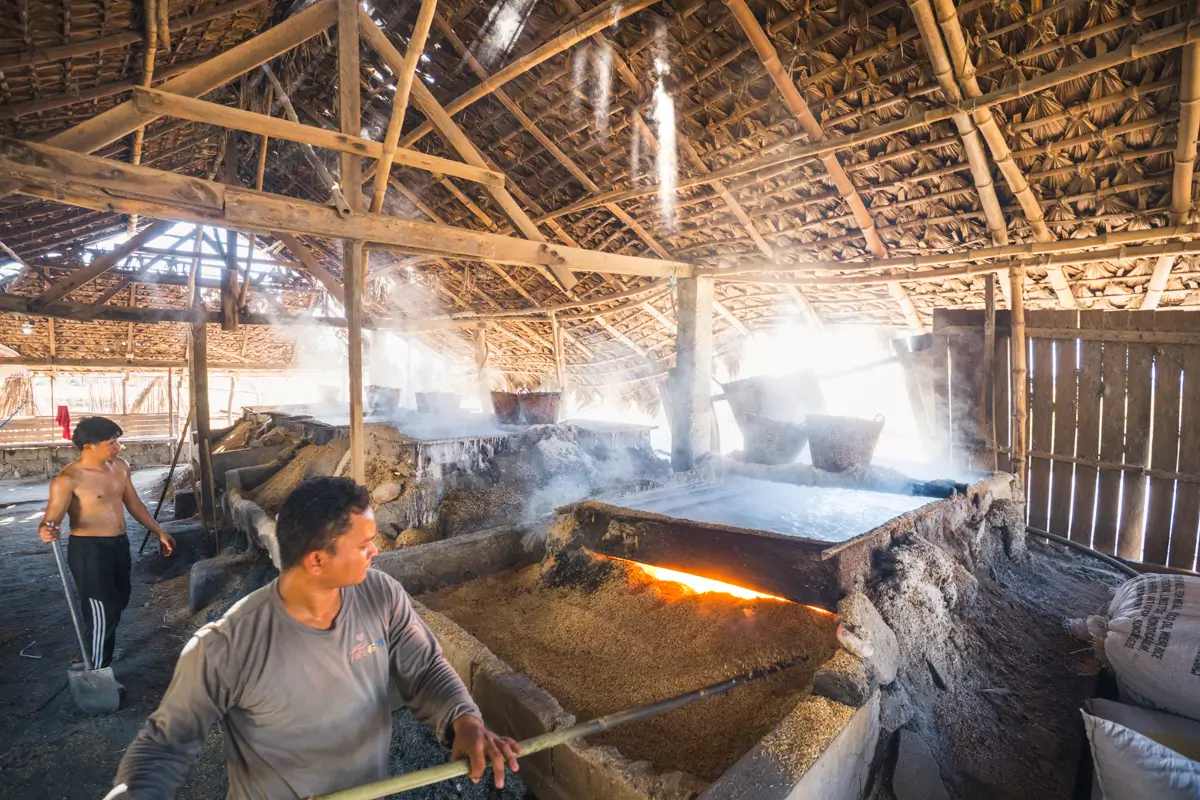 Two men work inside a rustic structure with a thatched roof, tending steaming vats over open flames—a glimpse into traditional crafts found at some of the best places in Vigan. Sunlight streams through roof gaps, adding charm to this unique Vigan tourist spot.