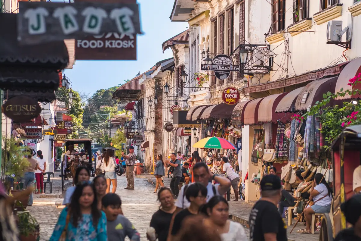 A busy cobblestone street lined with old-style buildings, shop signs, and awnings—one of the best places in Vigan. People walk, shop, and interact amid colorful displays and street vendors, creating a lively scene typical of famous Vigan tourist spots.