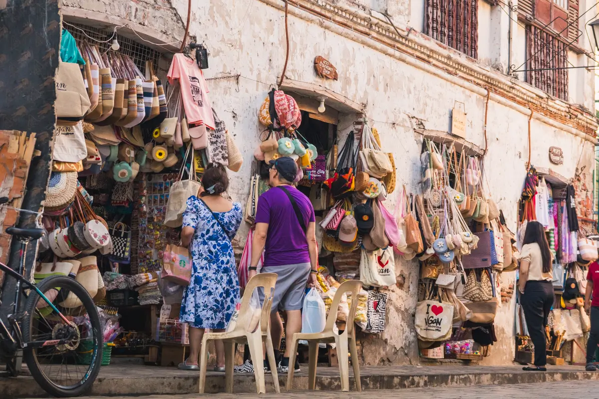Three people stand outside a street shop with a weathered wall, browsing bags and hats—a scene straight from any Vigan travel guide. A bicycle is parked on the left, with two plastic chairs near the shoppers exploring the best places in Vigan.
