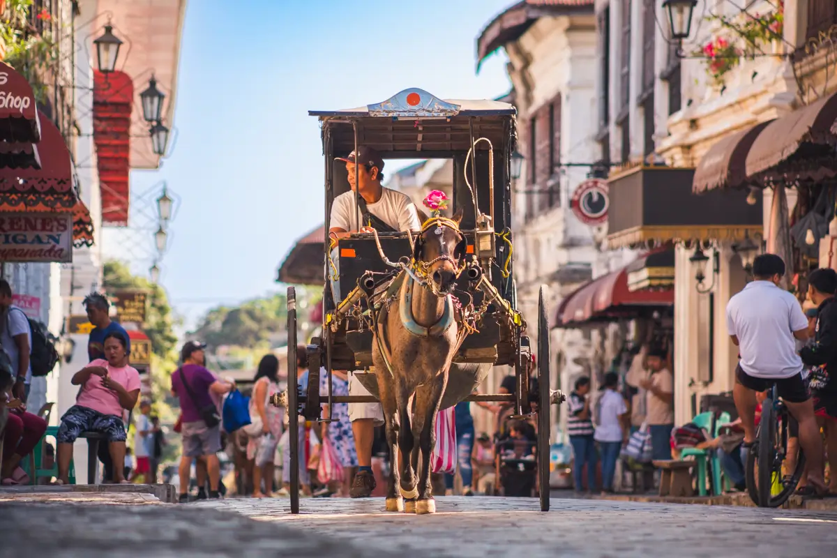 A horse-drawn carriage with a driver moves down a cobblestone street lined with people, shops, and historic buildings under daylight. Discover this vibrant scene among the best Vigan tourist spots in any Vigan guide.