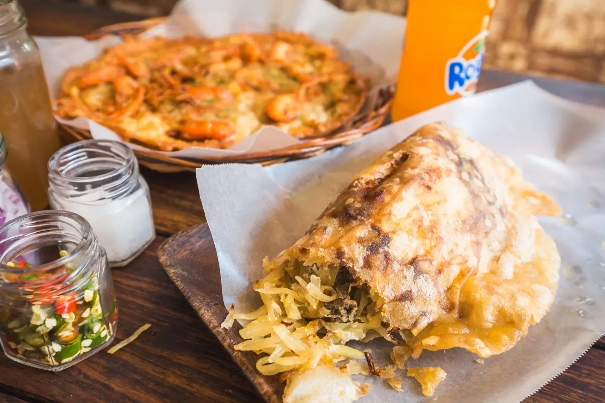 A fried, stuffed pancake filled with shredded vegetables sits on parchment paper. In the background are jars of condiments, a seafood-topped dish, and a bottle of orange soda—perfect fuel before exploring the best places in Vigan.