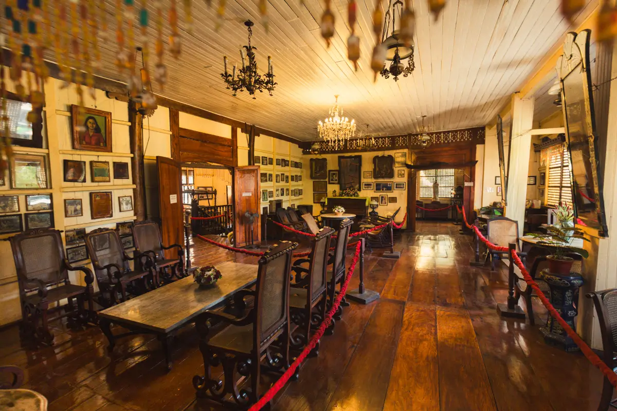 A spacious, historic room with polished wooden floors, antique chairs and tables, chandeliers, and walls decorated with framed photos—perfect for any Vigan travel guide exploring famed Vigan attractions. Red ropes section off areas as sunlight streams through the windows.