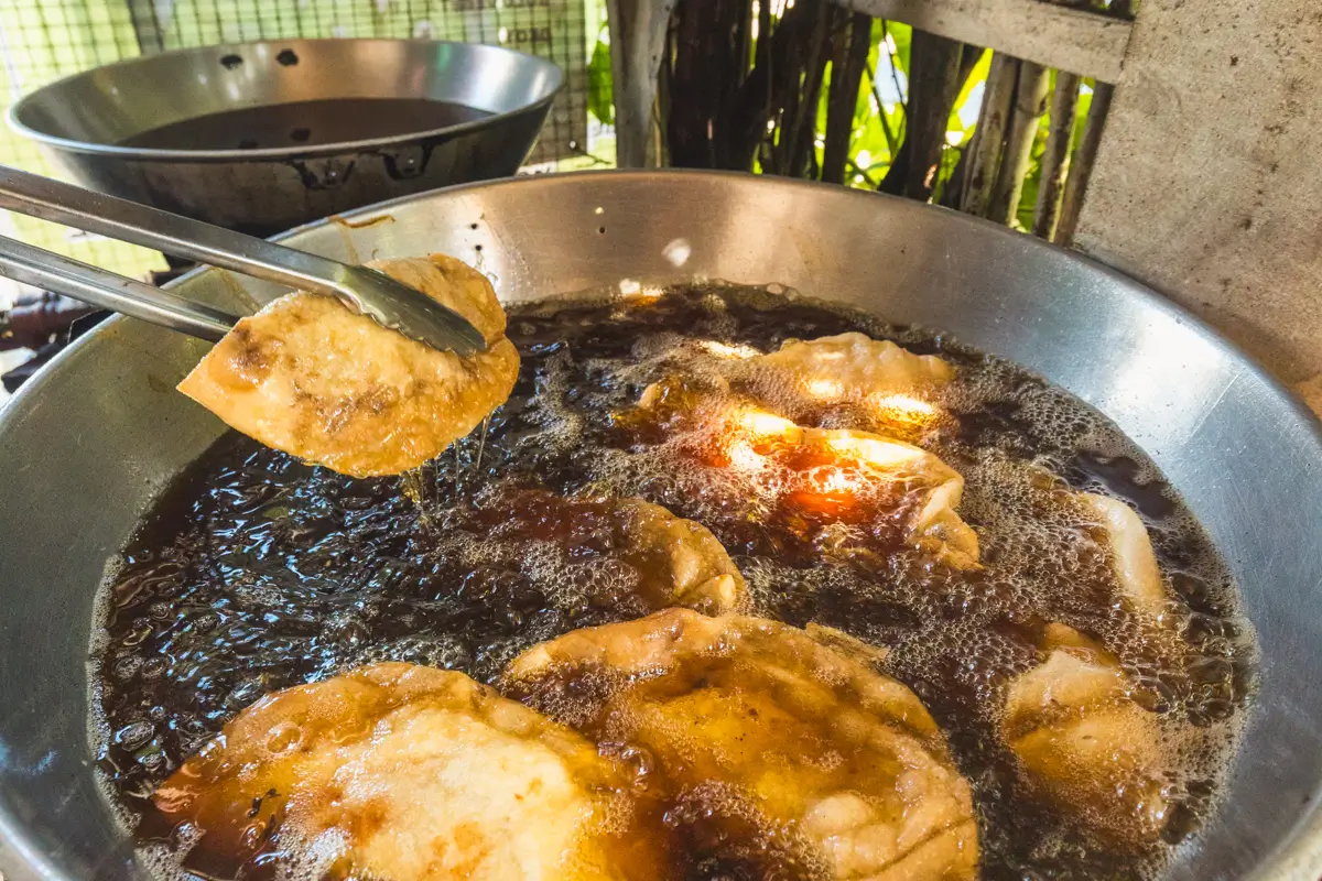 A metal pan filled with hot oil is frying dough pieces, a classic scene from the best places in Vigan. One piece is being lifted out with metal tongs, while another empty pan sits on the counter in the background.