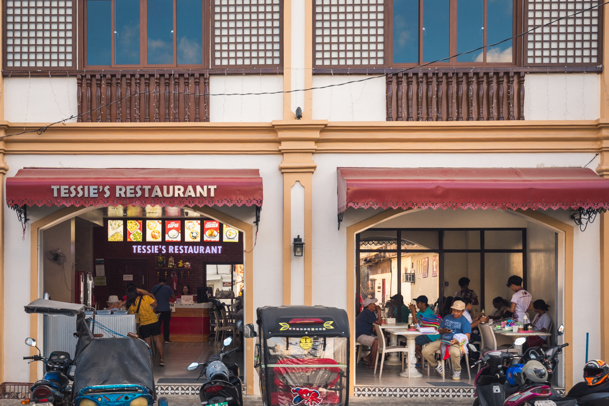 Street view of Tessie’s Restaurant, a popular stop featured in many Vigan travel guides. People dine in the open area while several motorcycles and tricycles are parked outside the cream and brown building.