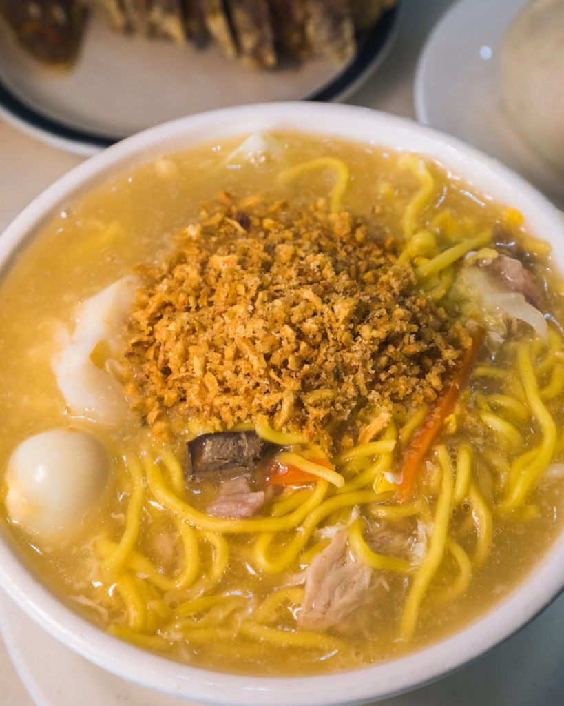 A bowl of noodle soup with yellow noodles, quail egg, vegetables, meat pieces, and a topping of fried garlic bits in a thick broth—just one of the local flavors you’ll find highlighted in any Vigan travel guide. Placed on a light-colored table.