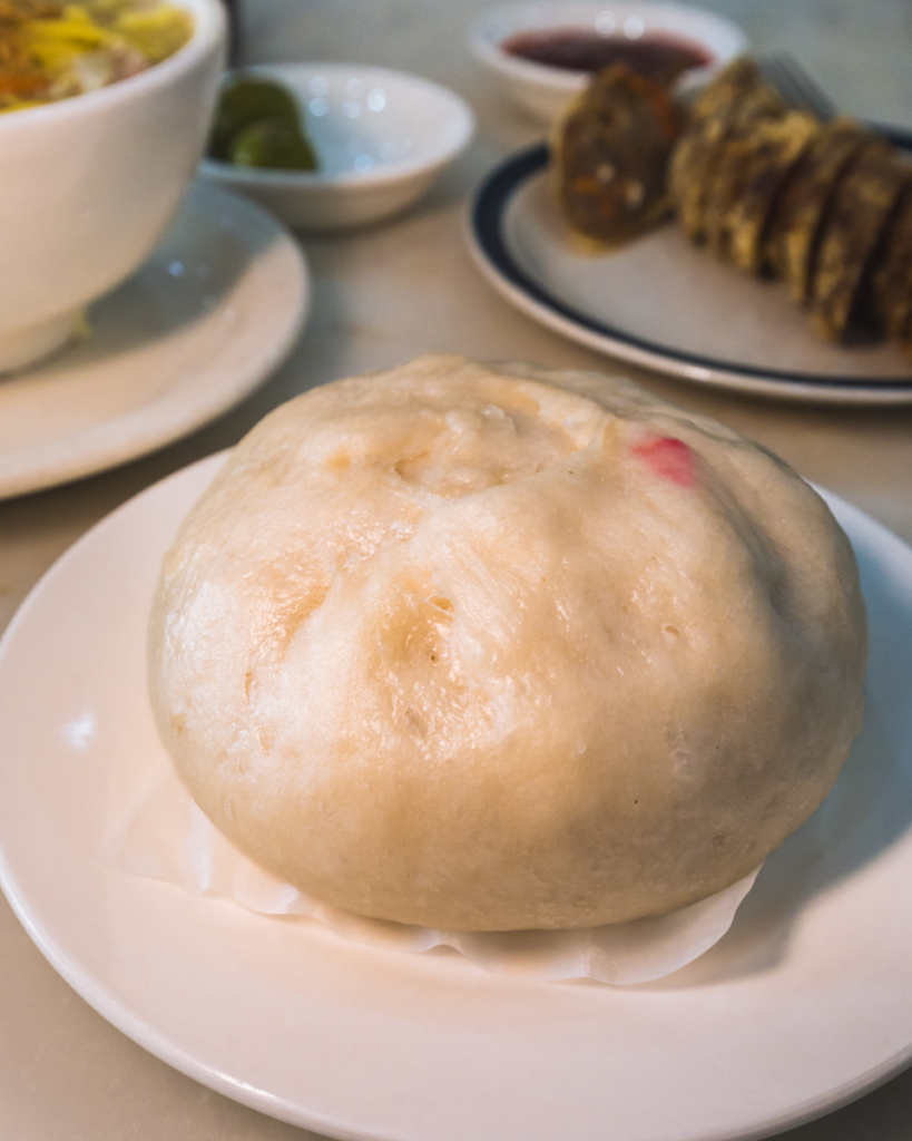 A large steamed bun sits on a white plate, with other dishes such as sliced food on another plate and a bowl partially visible in the background—a taste of local flavors you’ll find at the best Vigan tourist spots.