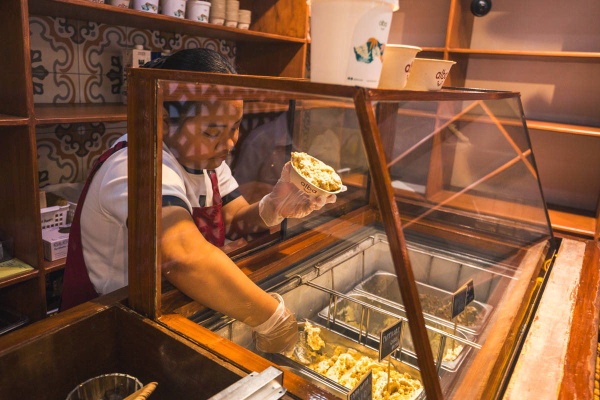 A person wearing gloves and an apron serves ice cream from a display case, holding a cup filled with ice cream—a delightful treat often enjoyed by visitors exploring famous Vigan attractions and other Vigan tourist spots.