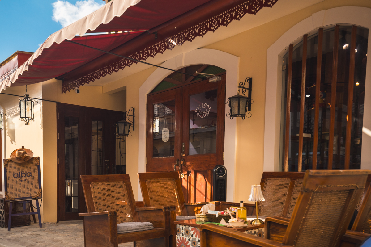 Outdoor seating area of a café with wooden chairs and a table, located in front of a beige building with large windows, a glass door, and a red awning—one of the best places in Vigan for visitors exploring top Vigan tourist spots.
