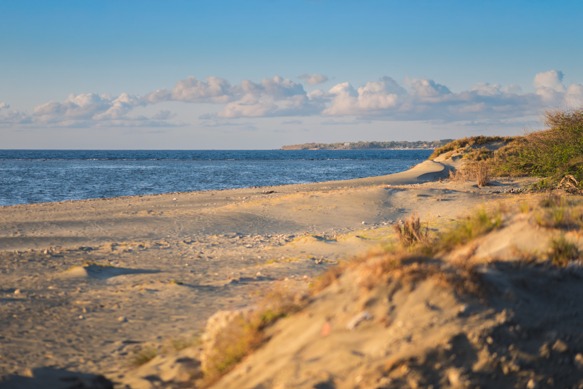 A sandy beach with scattered vegetation stretches along the shoreline beside calm blue water, resembling the serene coastal landscapes often featured in Vigan travel guide highlights. The coastline curves gently, with distant land visible on the horizon.