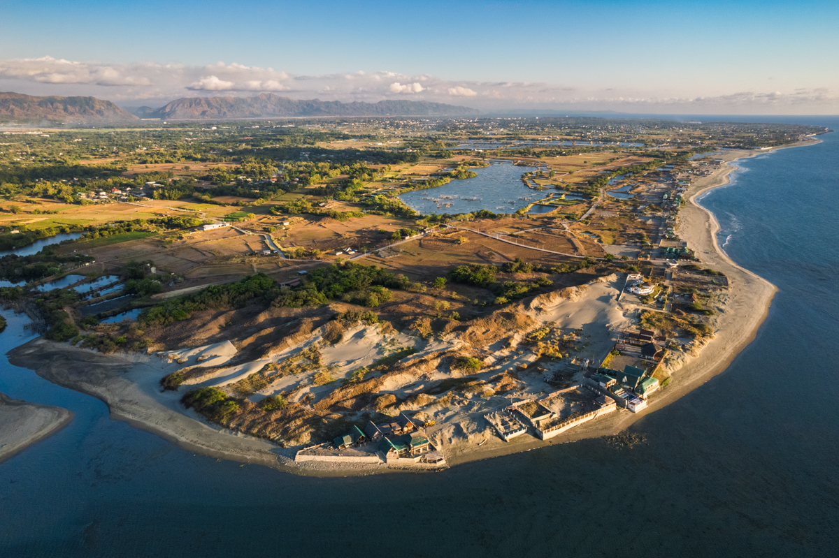 Aerial view of a coastal area with sandy dunes, shoreline buildings, a river mouth, and farmland extending inland—an inspiring scene for any Vigan travel guide highlighting the best things to do in Vigan and discover hidden tourist spots.