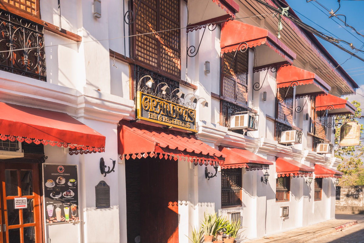 A white building with red awnings, barred windows, and air conditioning units; a sign reading Granpa’s Inn & Grill hangs above the entrance. Featured in many Vigan travel guide lists as one of the best places in Vigan for local cuisine.