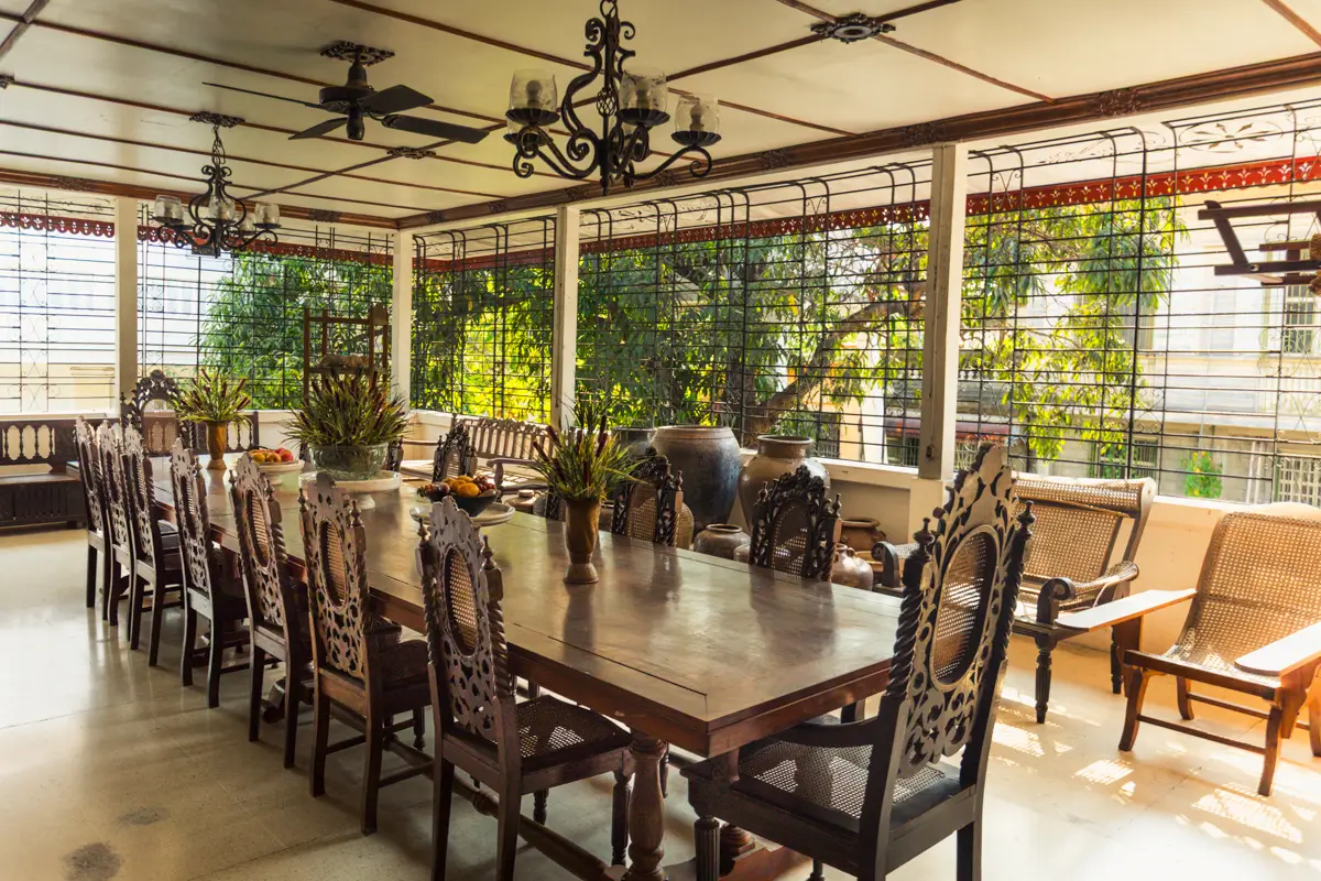 A large wooden dining table with ornate chairs sits on a covered patio surrounded by metal-grilled windows and lush greenery outside—evoking the timeless charm found in Vigan tourist spots. Ceiling fans and decorative lighting fixtures hang above.