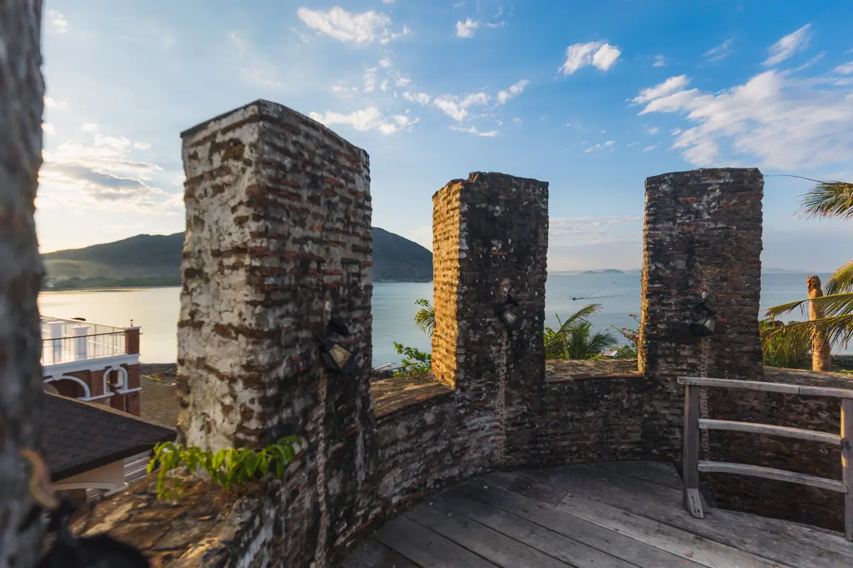Four weathered brick columns stand on a curved stone terrace overlooking calm water, with mountains and a blue sky beyond—a peaceful scene often featured in the Best Vigan guides for those seeking top Vigan tourist spots.