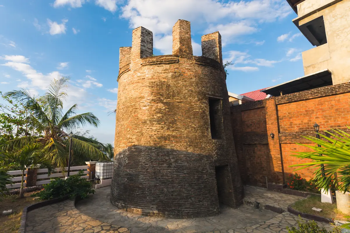A round brick tower with four battlements stands beside a brick wall and a modern building, surrounded by palm trees and paved stone ground under a partly cloudy sky—one of the best places in Vigan for your Vigan travel guide.