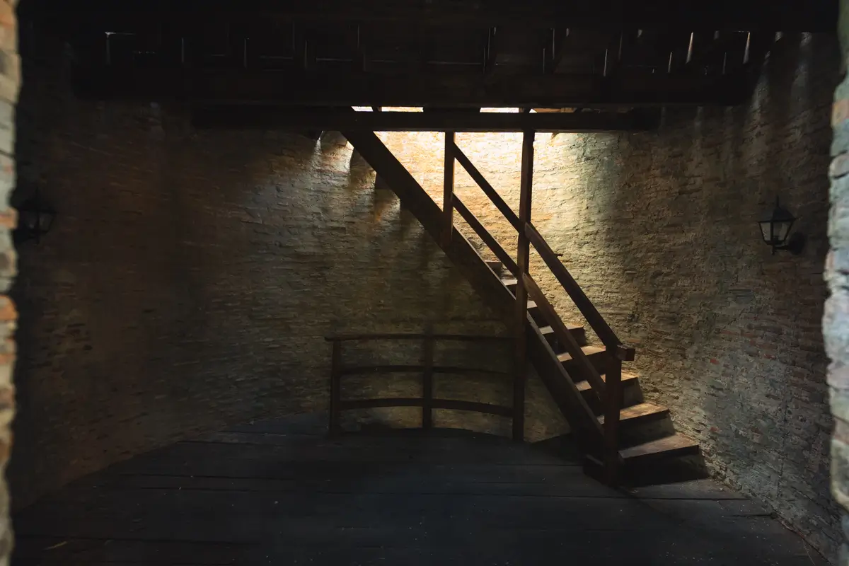 A wooden staircase leads up to a loft area in a dimly lit room with brick walls and a dark floor—reminiscent of the best Vigan interiors. Sunlight streams in from above, casting shadows beside two wall-mounted lamps.