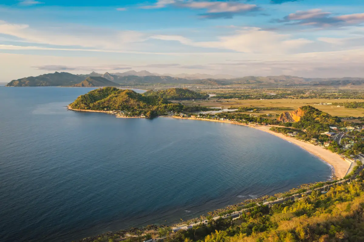 A wide bay with calm blue water curves along a sandy coastline bordered by green hills and small buildings, with distant mountains under a partly cloudy sky—one of the best places in Vigan featured in any Vigan travel guide.