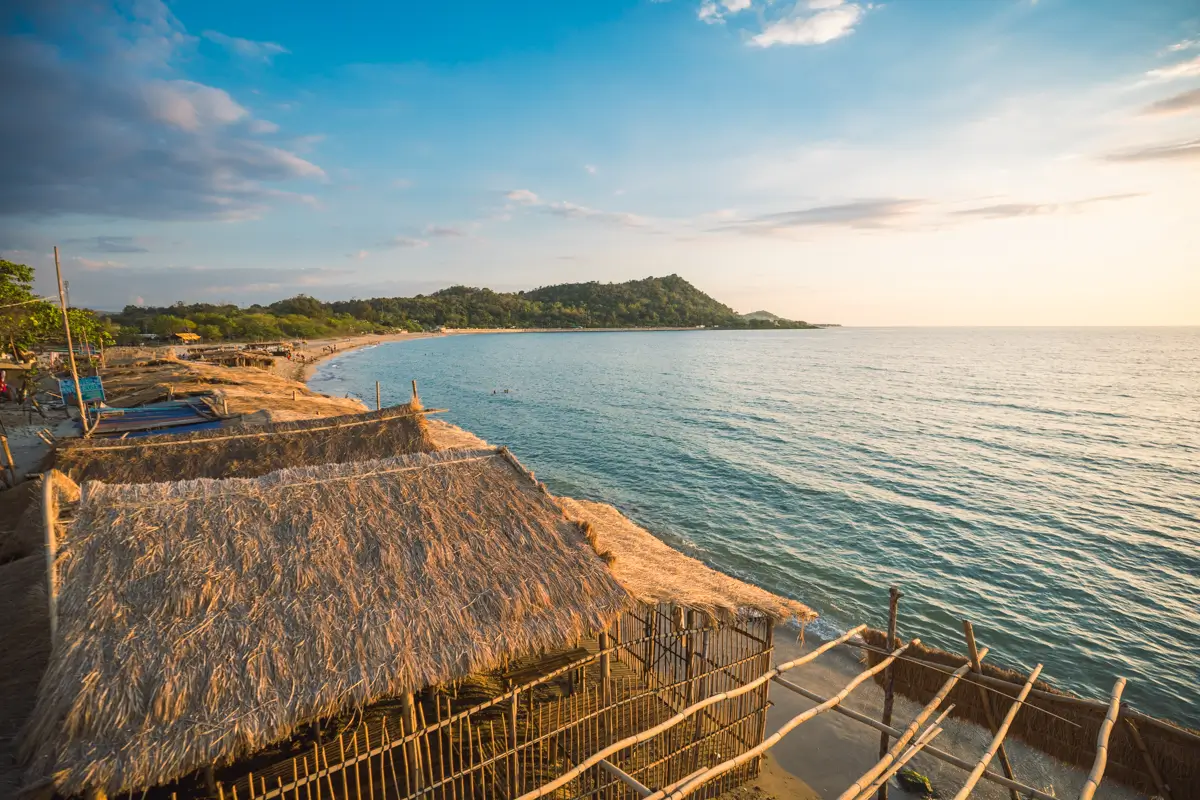 A coastal scene with thatched-roof huts near the shoreline, calm blue sea, and a distant green hill under a partly cloudy sky at sunset—an idyllic view often featured in the best Vigan attractions for any Vigan travel guide.