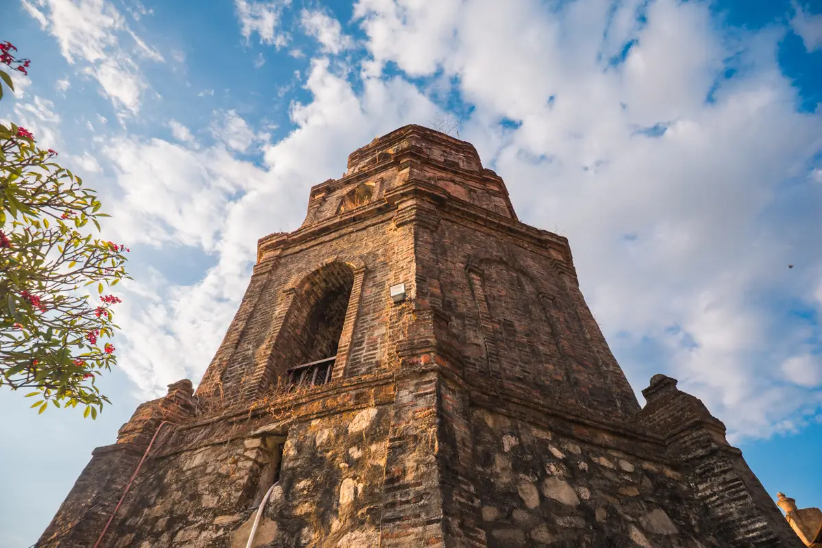 A low-angle view of an old brick and stone tower with weathered surfaces, set against a blue sky with scattered clouds—a scene straight from a Vigan travel guide. Some green foliage with red flowers appears on the left side of the image.