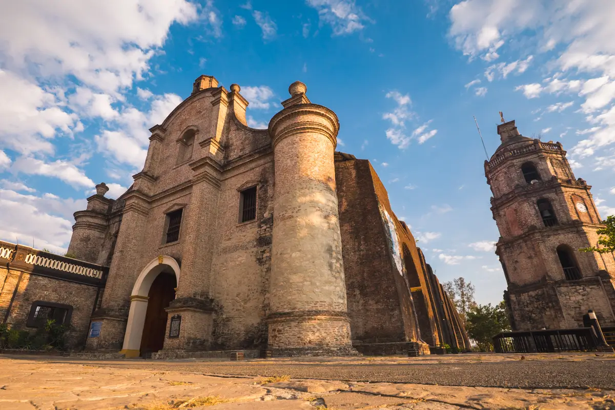 A historic stone church, featured in many Vigan travel guides, stands with two rounded columns at its entrance, an arched doorway, and a detached bell tower beneath a blue sky with scattered clouds. The photo is taken from a low angle.