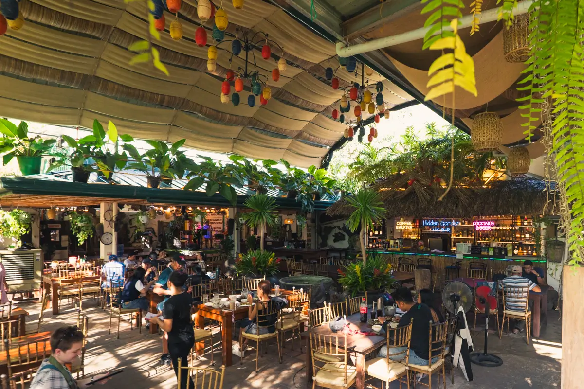 A covered outdoor restaurant with wooden tables and chairs, hanging plants, and ceiling decorations offers a cozy vibe. People are dining while a bar with neon signs glows in the background—one of the best Vigan tourist spots under a canopy roof.