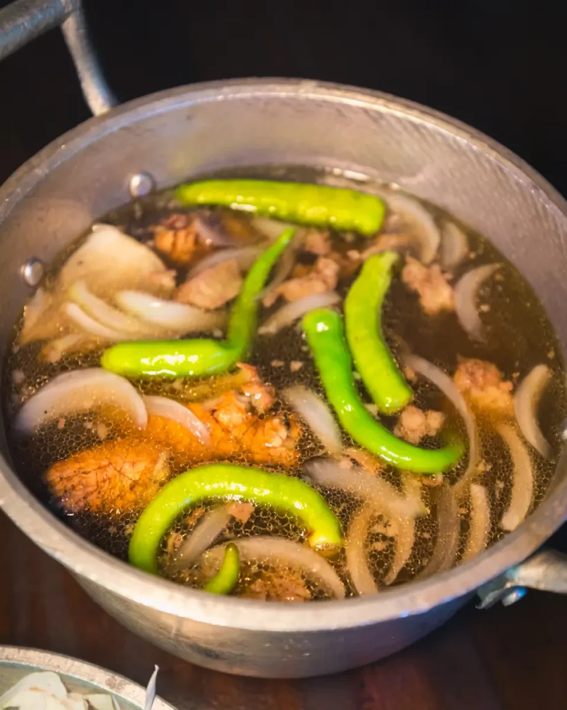 A metal pot filled with broth containing pieces of meat, sliced onions, and whole green chili peppers—just one of the local flavors you can savor when exploring the best places in Vigan featured in any Vigan travel guide.