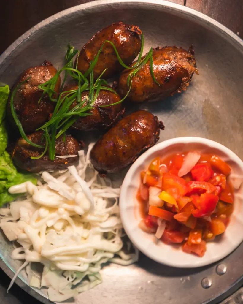 A metal plate with grilled sausages topped with green onions, shredded cabbage, lettuce, and a small bowl of chopped tomatoes and onions—a savory treat inspired by the best places in Vigan.