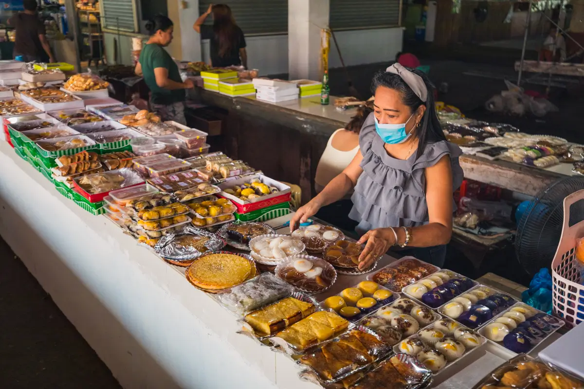 A woman wearing a face mask arranges various traditional Filipino desserts and sweets displayed in plastic containers on a market stall table—a vibrant scene often highlighted in Vigan tourism guides. Other people and stalls are visible in the background.