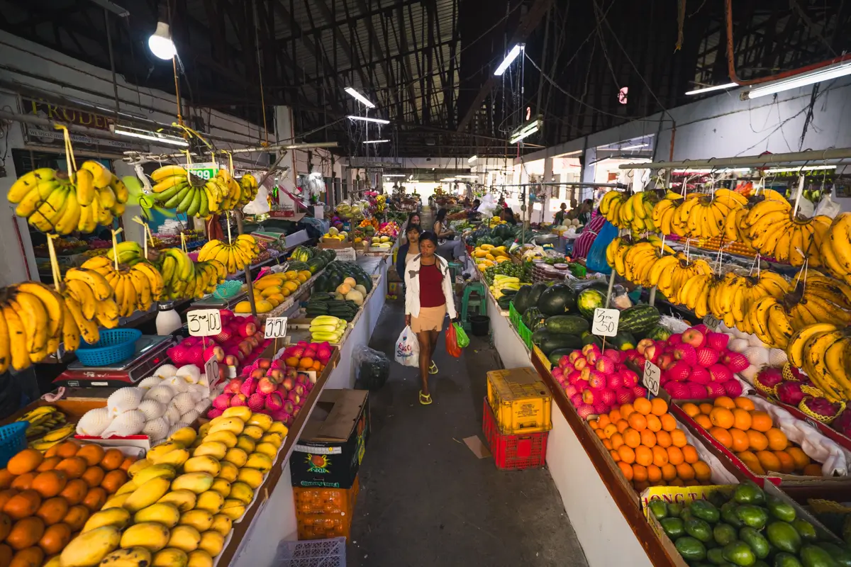 A person walks through an indoor market, one of the vibrant Vigan tourist spots, lined with colorful fruit stalls displaying bananas, papayas, dragon fruit, and oranges under a high roof with hanging lights.