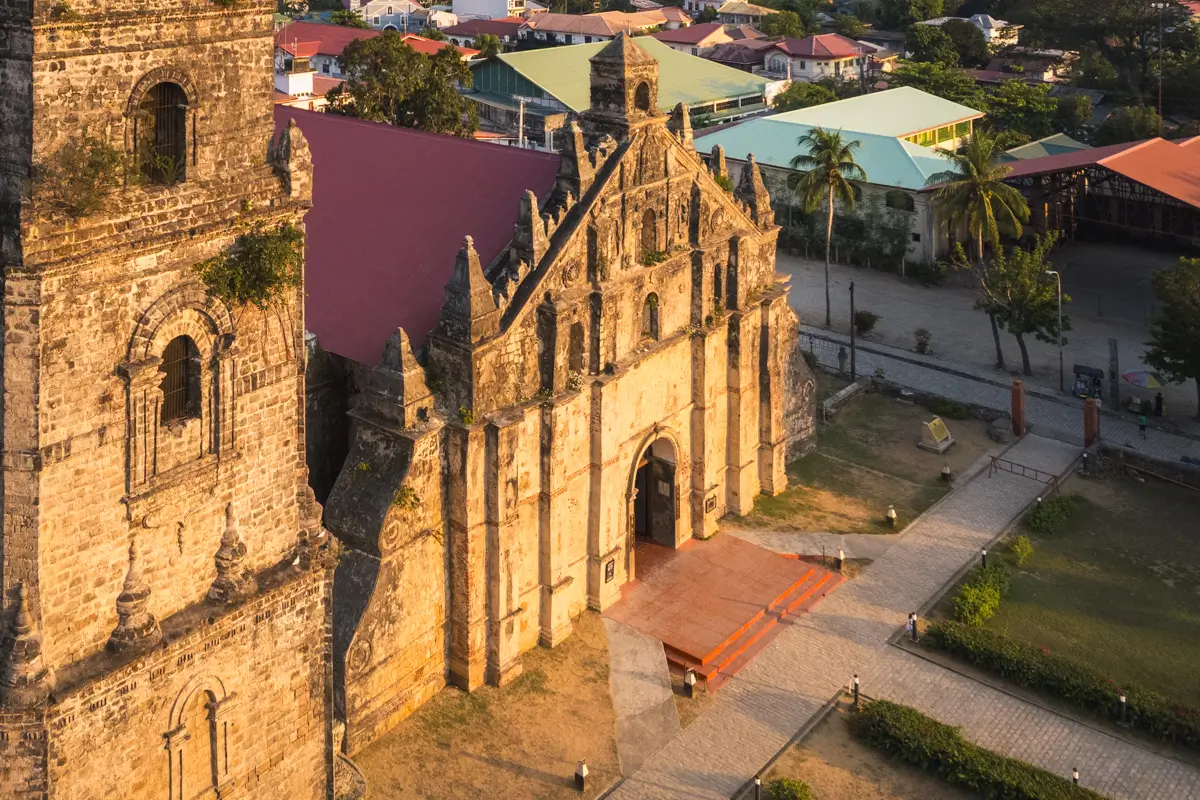 Aerial view of a historic stone church with a tall bell tower, arched entrance, and gabled façade—one of the best Vigan tourist spots—surrounded by a plaza, pathways, and colorful residential roofs.