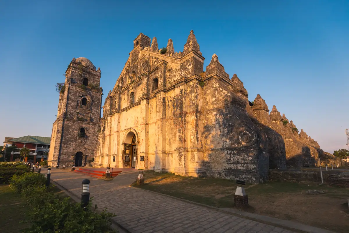 A stone church with a prominent bell tower, buttresses, and weathered facade stands under a clear blue sky, surrounded by greenery—a must-see among Vigan tourist spots, beautifully illuminated in the late afternoon.