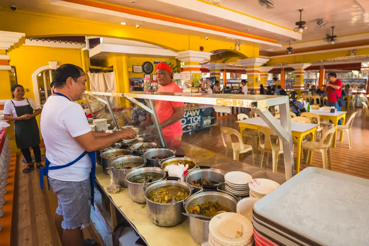 A man wearing an apron serves food from metal pots behind a glass counter in a brightly lit cafeteria—one of the best places in Vigan to stop by after exploring famous Vigan tourist spots, with yellow chairs and tables filling the background.