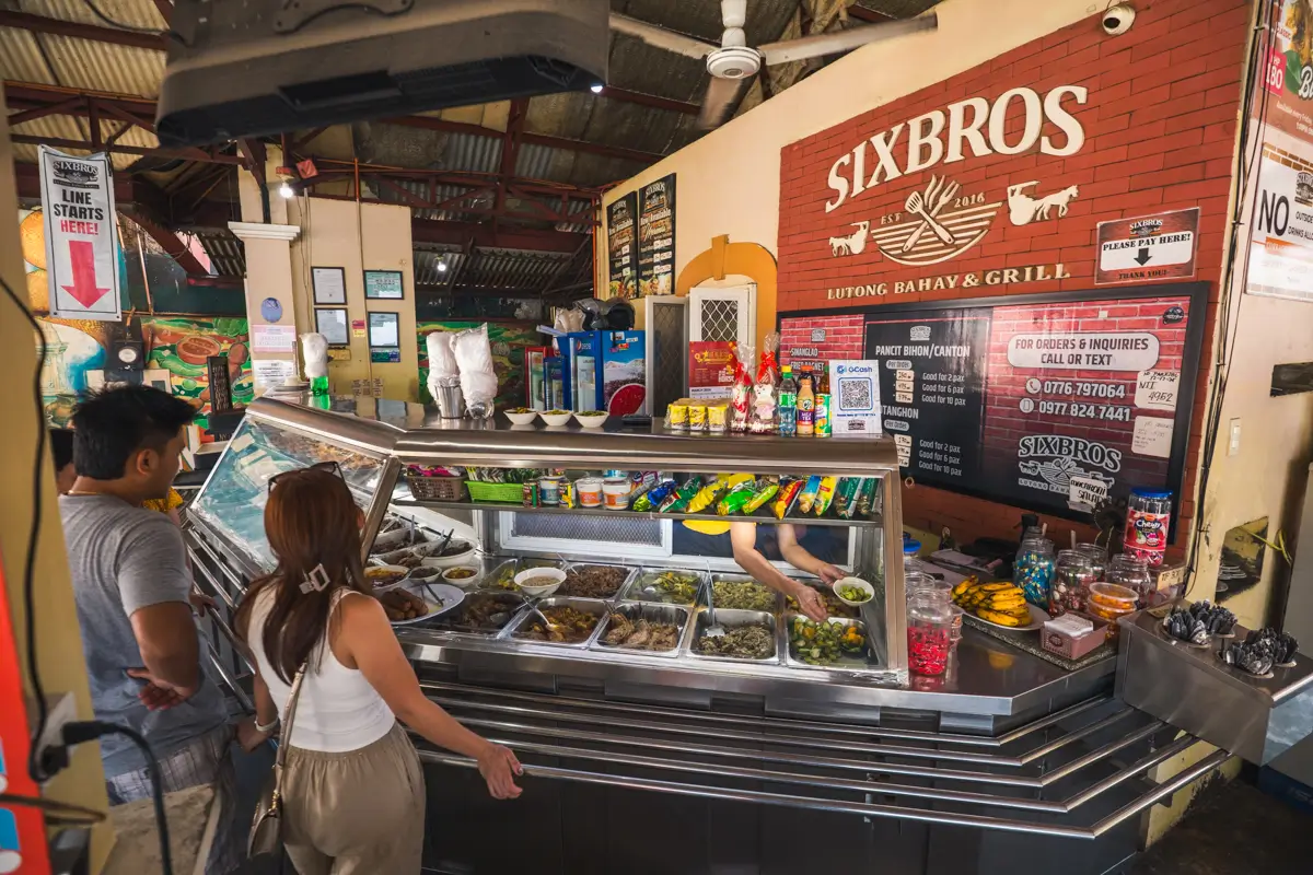 Two people stand at a food counter displaying various dishes in metal trays at Sixbros Hutong Bahay & Grill, a tasty stop often featured in Vigan guides for those exploring top Vigan attractions and local flavors.