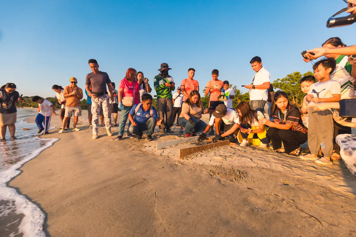 A group of people, including adults and children, are gathered on a sandy beach near the shoreline—one of the best places in Vigan—observing and photographing something on the ground as the sun sets.
