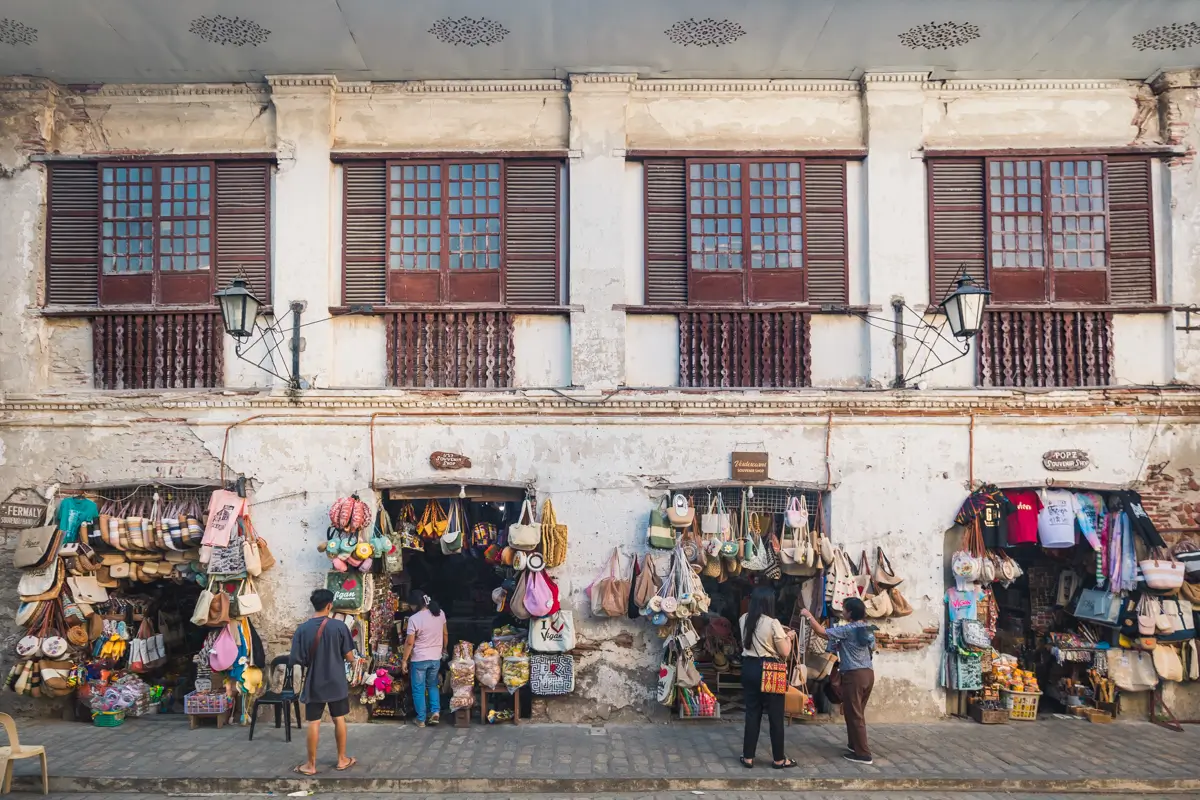 Street vendors sell bags and handicrafts displayed on a weathered building with wooden shutters. Several people browse the items along a cobblestone sidewalk—one of the best places in Vigan for unique finds and local charm.