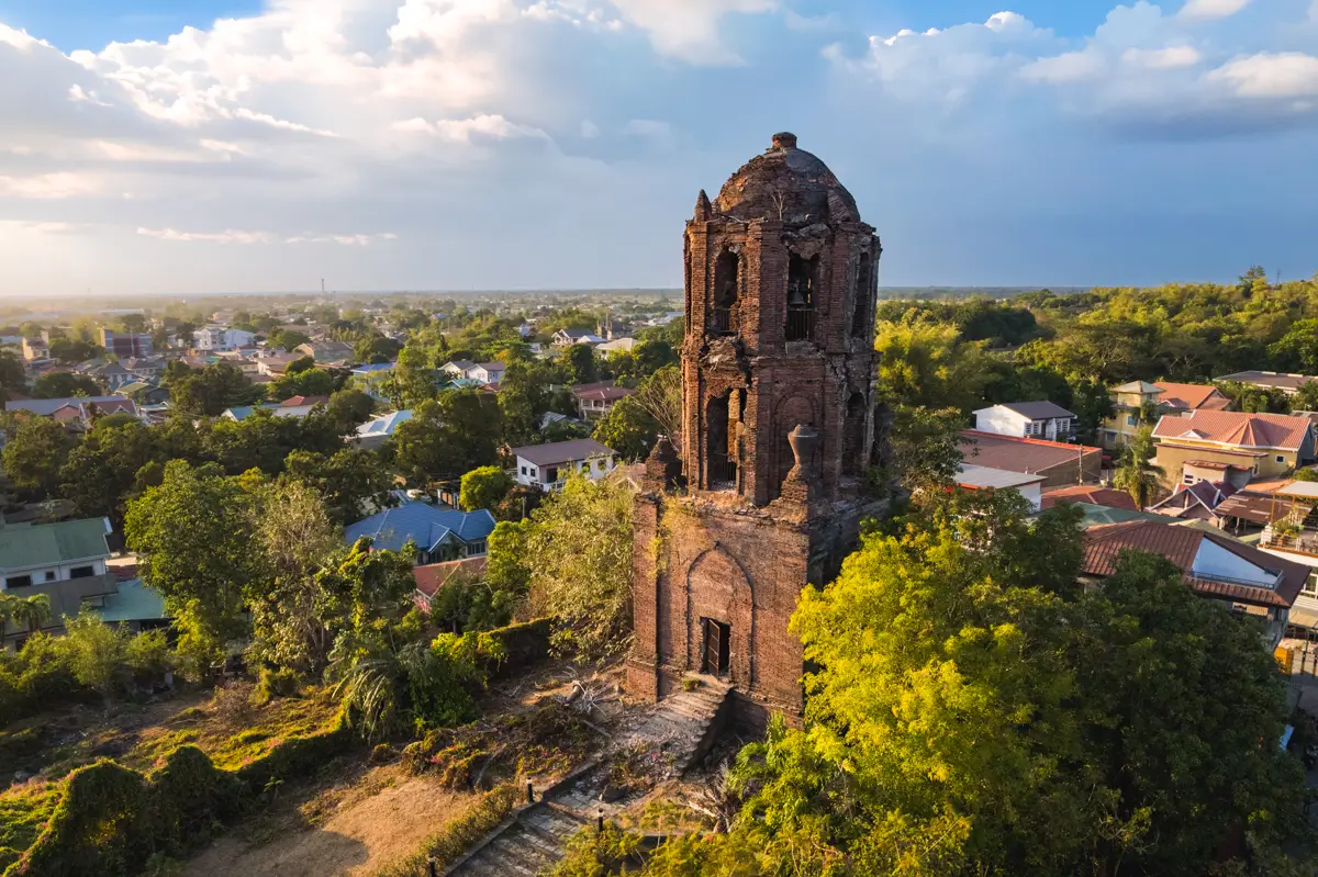 Aerial view of an old brick bell tower with arched windows standing on a hill surrounded by trees and residential houses under a partly cloudy sky—one of the best places in Vigan featured in many Vigan travel guide recommendations.