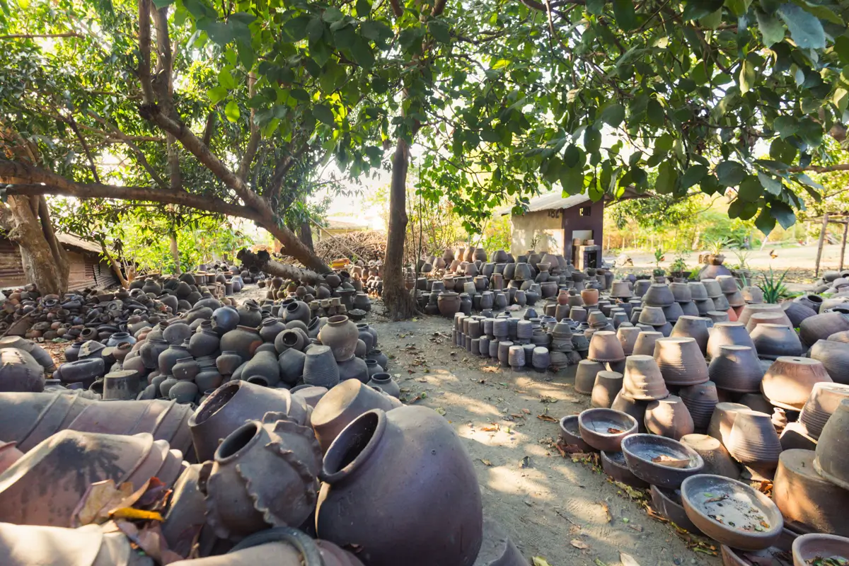 Hundreds of clay pots and vases are stacked on the ground under the shade of large trees in an outdoor pottery workshop, one of the unique Vigan tourist spots. Sunlight filters through the leaves, illuminating parts of the pottery and workshop area.